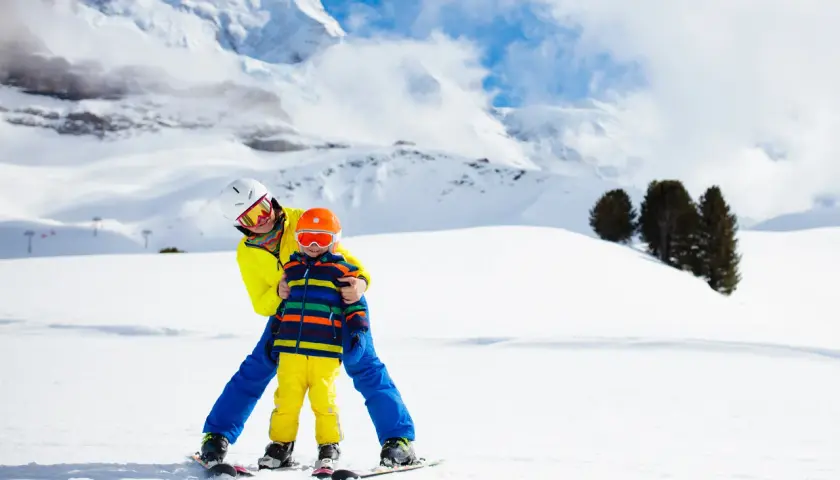 Un adulto insegna a sciare a un bambino di 2 anni su una pista innevata, con montagne sullo sfondo. Un perfetto weekend con bambini 2 anni!