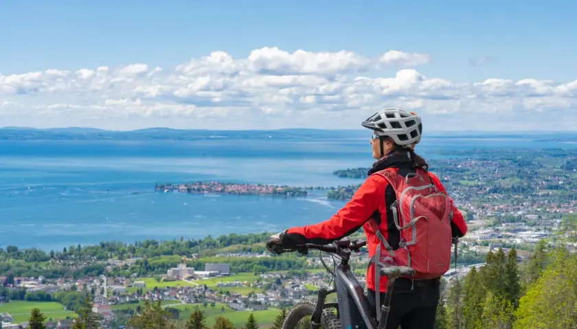 A woman in a red jacket and helmet pauses her **Lake Constance bike tour** to admire the expansive blue lake and distant towns under a cloudy sky.