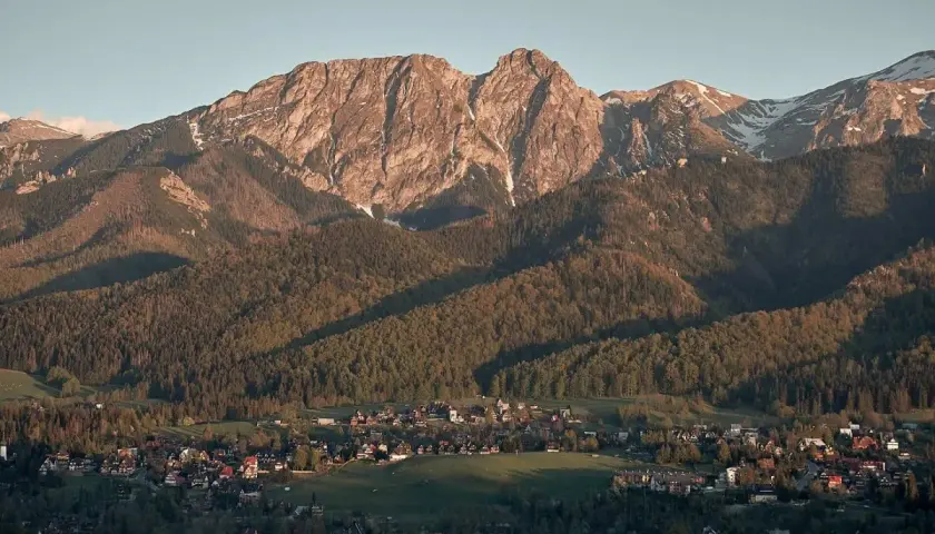 Widok z **Antałówki punkt widokowy** na Tatry. Słońce oświetla skaliste szczyty i lasy, a w dole widać zabudowania.