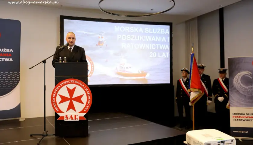 Mężczyzna przemawia przy mównicy z logo SAR. Na ekranie morska służba poszukiwania i ratownictwa.