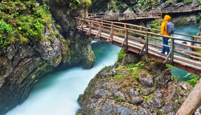 Ein Wanderer auf einer Holzbrücke über einem türkisfarbenen Fluss in einer Schlucht, ähnlich dem Bleder See.