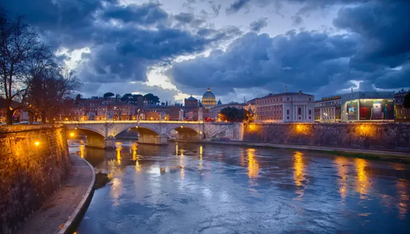 Il Tevere riflette le luci di Roma al tramonto, con la cupola di San Pietro sullo sfondo. La **Trastevere nightlife** è già nell'aria, promessa di serate vivaci.