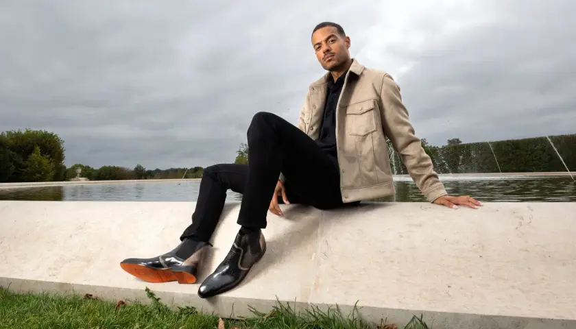 Un homme élégant montre comment porter des chelsea boots homme avec un jean noir et une veste beige, près d'une fontaine.