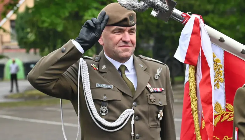 Pułkownik Suliwiński w mundurze salutuje obok polskiej flagi.
