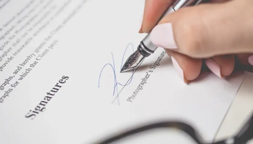 Mano con pluma estilográfica firma un documento. La firma se realiza encima o debajo del nombre "Photographer's Signature".