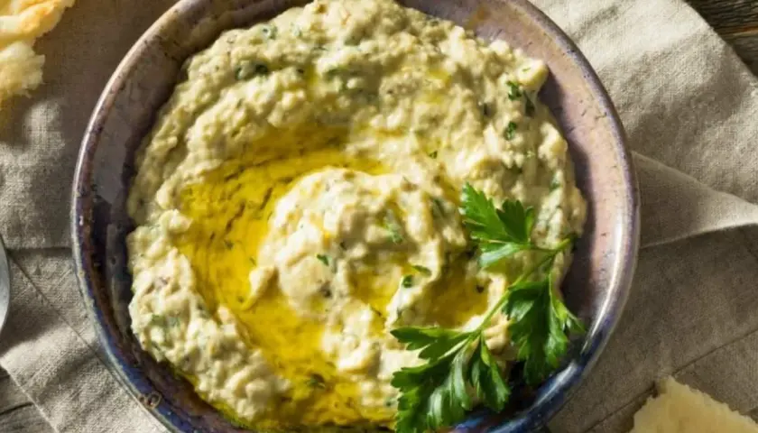 Baba ganush cremoso en un cuenco, con aceite de oliva y perejil fresco. Pan de pita al lado.