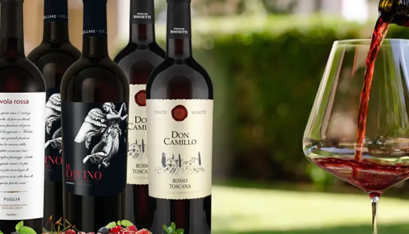 Flaschen mit teurem italienischem Rotwein, darunter "Il Divino" und "Don Camillo", werden vor einem Glas Rotwein präsentiert, in das gerade eingeschenkt wird. Frische Beeren und Kirschen liegen im Vordergrund.