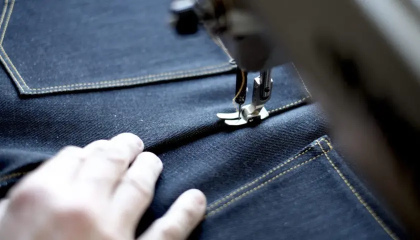 A sewing machine stitches a precise jean seam on dark denim, with a hand guiding the fabric.