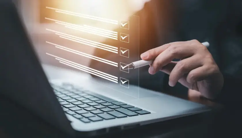Dłoń z długopisem zaznacza pozycje na liście kontrolnej wyświetlanej nad klawiaturą laptopa.