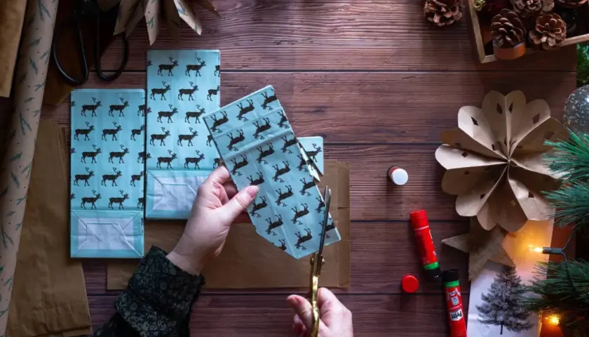 Ręce tną papier z reniferami, tworząc ozdoby choinkowe z papieru. Obok kleje, szyszki i choinka.