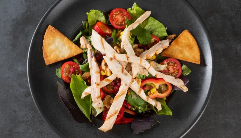Deliziosa insalata di pollo fredda con pomodorini, mais, peperoni e crostini croccanti su un piatto scuro.