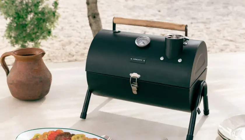 Barbecue noir avec couvercle, thermomètre et poignée en bois, prêt pour le temps de cuisson. Une carafe en terre cuite et une assiette de tomates colorées sont à proximité.