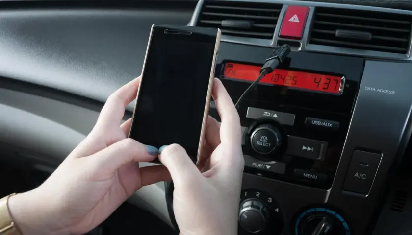 Ręce trzymają telefon, który jest podłączony kablem do radia samochodowego. Prosty sposób, jak połączyć telefon z samochodem.