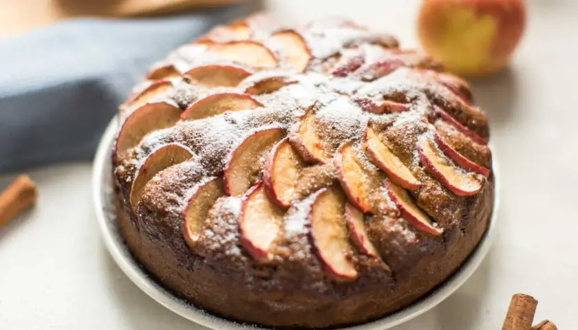 Un délicieux moelleux aux pommes, saupoudré de sucre glace, avec des pommes fraîches et des bâtons de cannelle en arrière-plan.