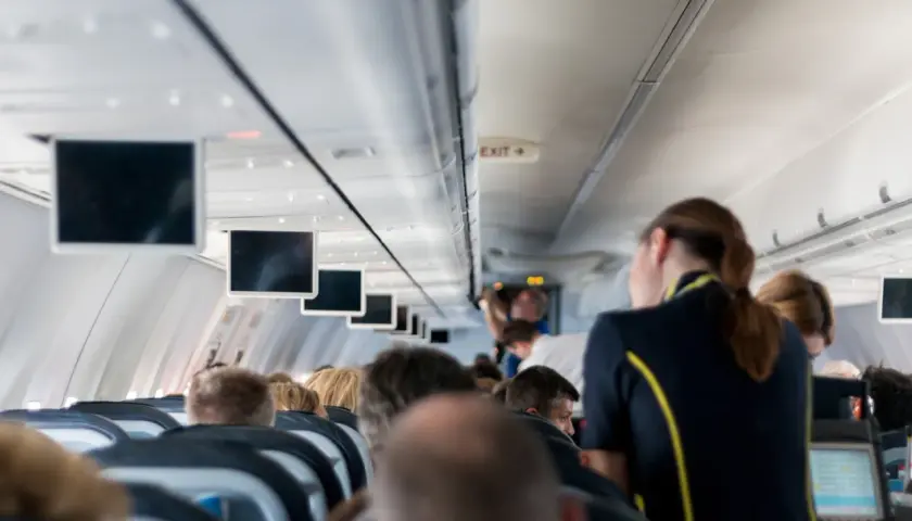 Widok z kabiny samolotu. Stewardka w granatowym mundurku z żółtym pasem obsługuje pasażerów.