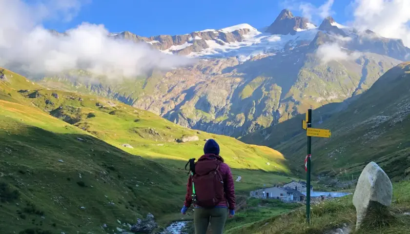 A hiker pauses on a **Tour Mont Blanc hike**, gazing at a majestic snow-capped mountain range. A signpost points the way to the next stage of this epic journey.