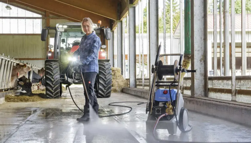 Frau reinigt Stallboden mit einem Industrie Hochdruckreiniger. Kühe und Traktor im Hintergrund.