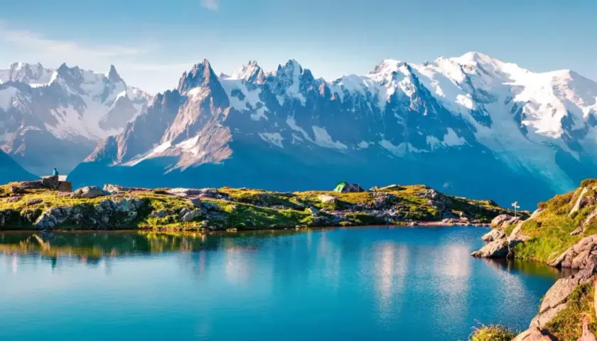 Un lago azul refleja la majestuosa **montaña Mont Blanc** y sus picos nevados. Un campista contempla el paisaje.