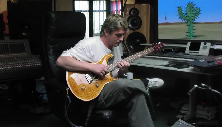 Mike Oldfield w studiu, gra na gitarze. Na ekranie komputerowym kaktus na pustyni, jak w jego utworach.
