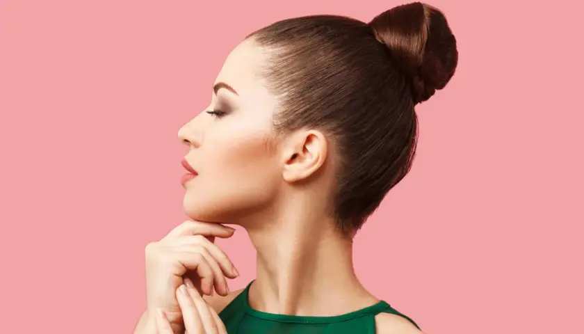 Mujer con moños altos elegantes, perfil sereno, mano en la barbilla, fondo rosa.