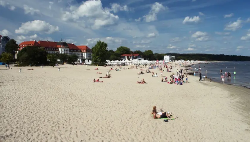 Plaża w Sopocie z ludźmi opalającymi się i spacerującymi, w tle budynki i morze.