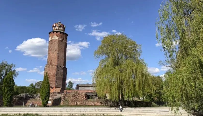 Wieża w Brodnicy Górnej, otoczona zielenią i ruinami, pod błękitnym niebem z chmurami.