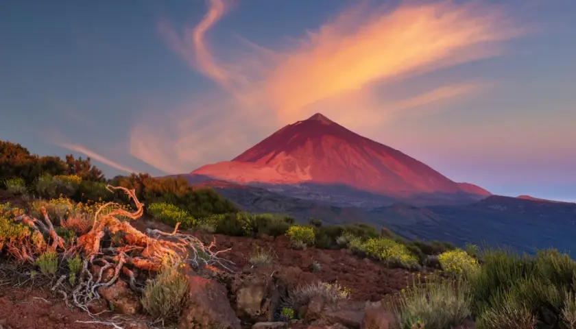 Volcán Teide bañado por el atardecer, ideal para actividades de montaña. Nubes anaranjadas y vegetación árida en primer plano.