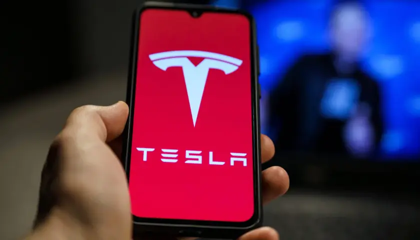 Dłoń trzyma telefon z logo Tesla. W tle widać niewyraźną postać na niebieskim tle, być może Elon Musk.