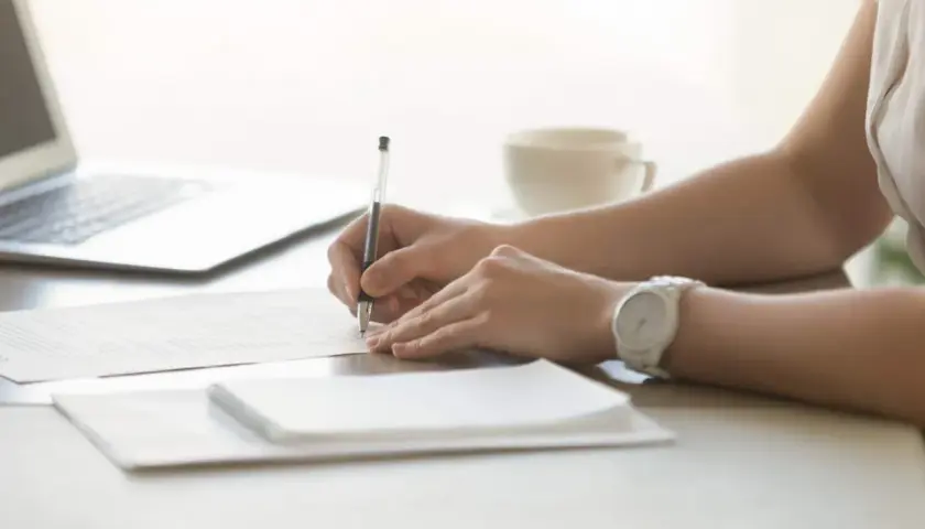 Ręce piszą dokument, być może oświadczenie najemcy zawierające wskazanie innego lokalu. Obok laptop i filiżanka kawy.