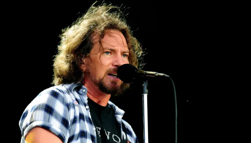 Eddie Vedder, wokalista Pearl Jam, śpiewa do mikrofonu na koncercie. Jego rozwichrzone włosy i charakterystyczna koszula dodają mu rockowego stylu.