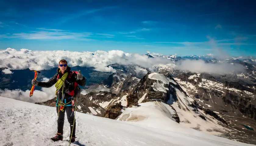 Alpinista en la cima del Gran Paradiso, con piolet y equipo, disfrutando de vistas espectaculares de montañas nevadas y nubes.