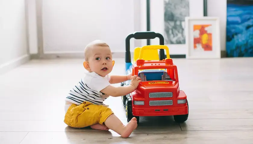 Uśmiechnięte niemowlę w żółtych spodenkach bawi się czerwono-żółtą ciężarówką na drewnianej podłodze.