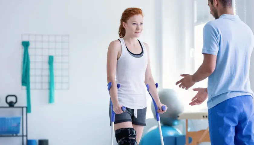 Kobieta na kulach z ortezą na nodze podczas zabiegów rehabilitacyjnych, wspierana przez fizjoterapeutę.