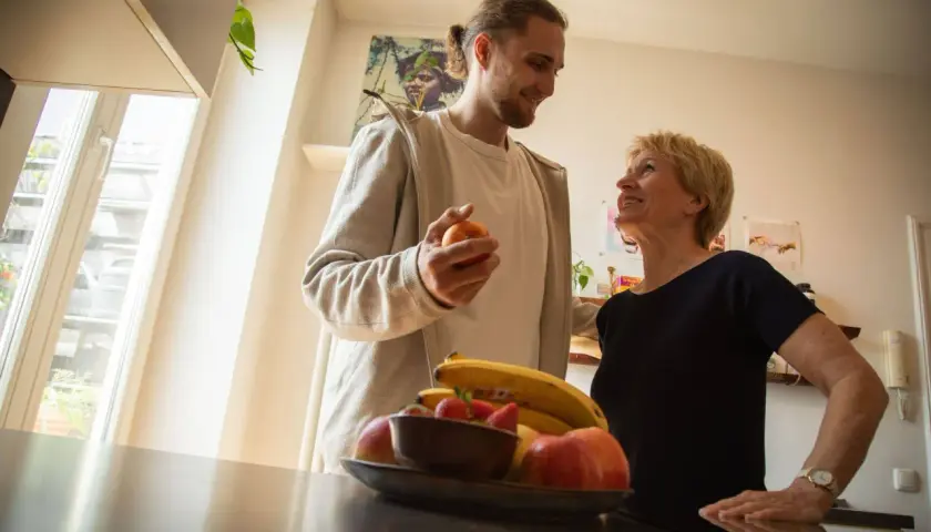 Ein Mann hält einen Apfel, während er mit einer lächelnden Frau spricht. Obst liegt auf einem Tisch. Vielleicht ein Moment der Entlastung für eine Privatperson.