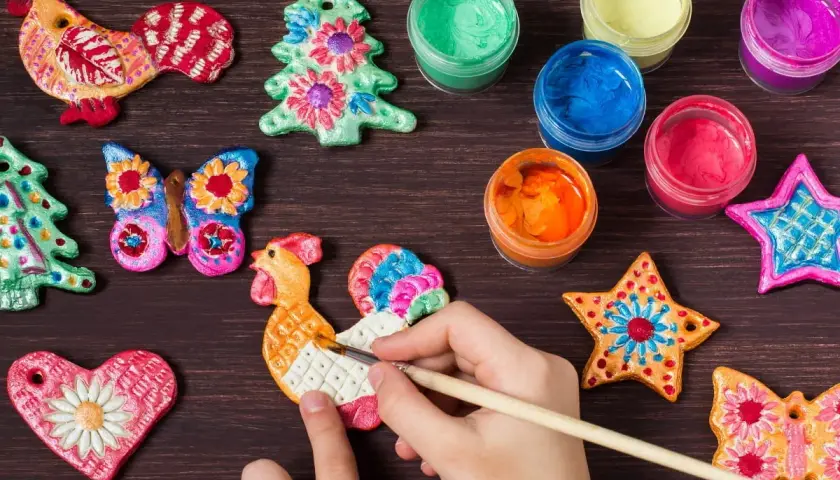 Dziecko maluje kurę z masy solnej, tworząc kolorowe inspiracje. Wokół ozdoby: choinki, motyle, serca, gwiazdki i farby.