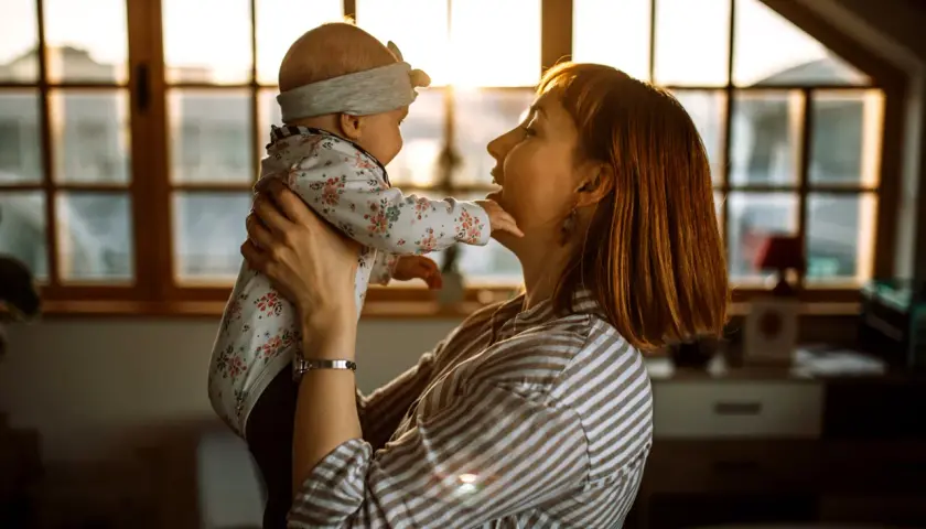 Mama bawi się z dzieckiem, które wykonuje odginanie głowy do tyłu. Ciepłe światło zachodzącego słońca oświetla ich twarze.