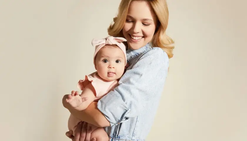 Uśmiechnięta mama tuli niemowlę w różowym body i opasce. Wskazówka, jak ubierać noworodka jesienią, by było mu ciepło i wygodnie.