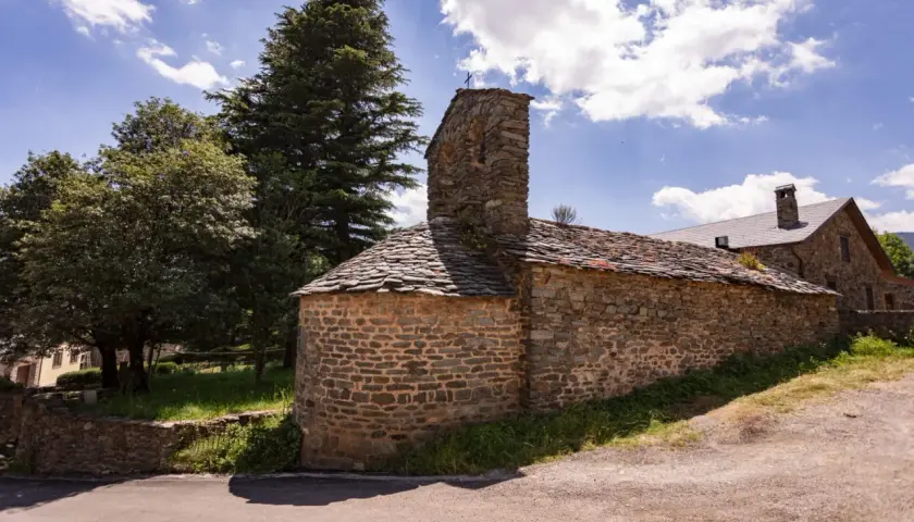 Iglesia románica de piedra con tejado de pizarra en el Ripollès, bajo un cielo azul con nubes algodonosas.
