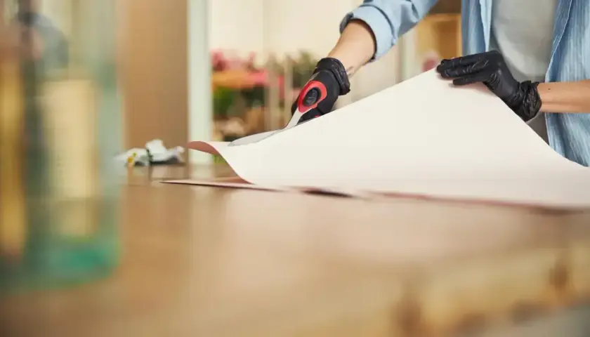 Osoba w rękawiczkach tnie papier, przygotowując go do przyklejenia. Zastanawiasz się, czym podkleić tapetę?