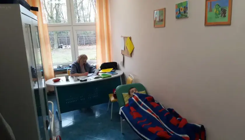 Pielęgniarka szkolna przygotowuje materiały do kursu online, podczas gdy dziecko odpoczywa pod kocem.