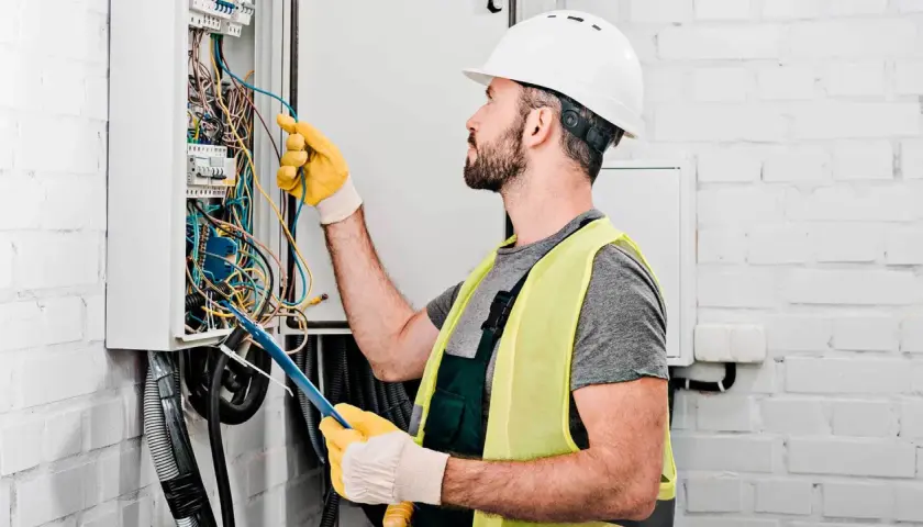 Elektryk w kasku i kamizelce odblaskowej pracuje przy skrzynce z przewodami. Zastanawiasz się, ile kosztuje elektryk?
