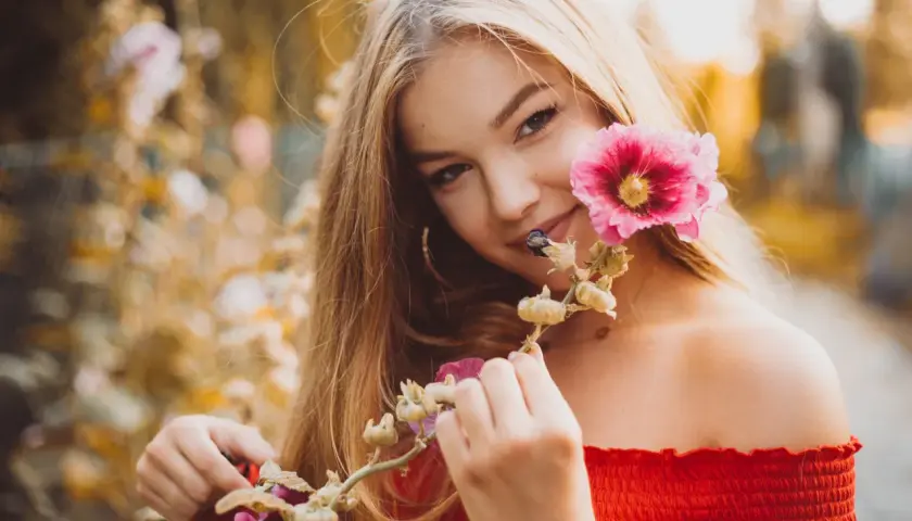 Uśmiechnięta blondynka w czerwonej bluzce podczas sesji kobiecej w plenerze, trzymająca gałązkę z różowymi kwiatami malwy.