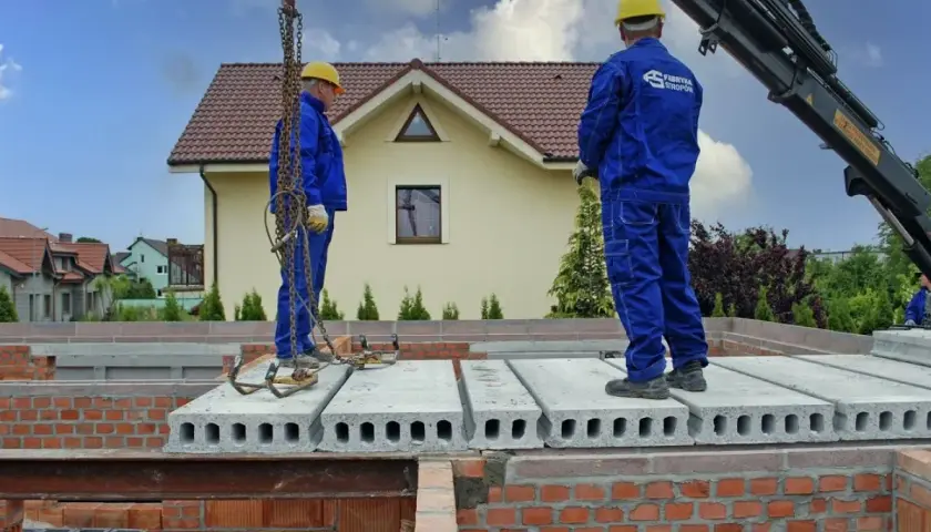 Dwóch pracowników w niebieskich kombinezonach montuje prefabrykowany strop. Dźwig podnosi kolejne płyty stropowe.