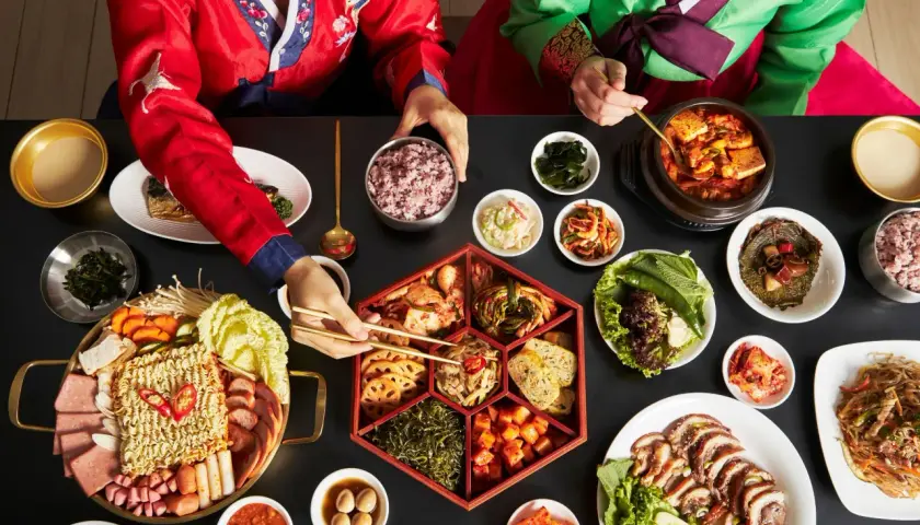 Dwie osoby w tradycyjnych koreańskich strojach jedzą obfity posiłek z wieloma daniami, w tym kimchi.