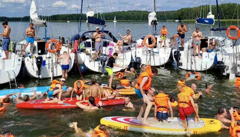 Ludzie w kamizelkach ratunkowych i strojach kąpielowych pływają i bawią się na deskach wodnych przy żaglówkach. Idealny długi weekend czerwiec na wodzie!