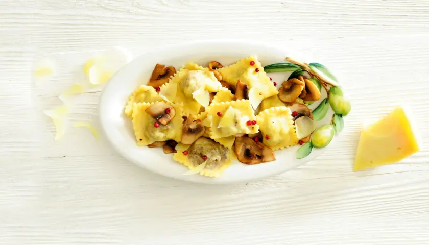 Pyszne ravioli z grzybami, posypane parmezanem i różowym pieprzem, podane na talerzu z gałązką oliwną.