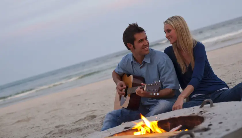 Coppia sulla spiaggia al tramonto, lui suona la chitarra, lei lo guarda sorridendo. Un falò illumina la scena, ma la noia nella coppia è palpabile.