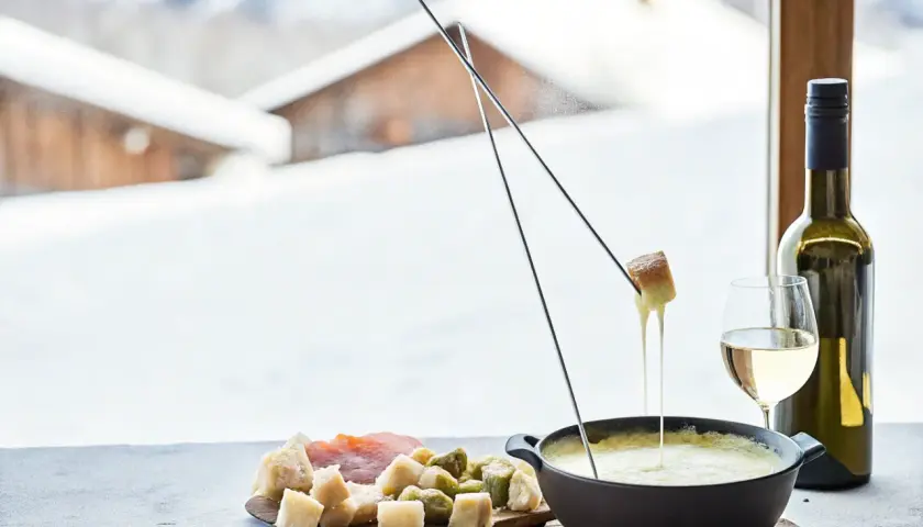 Une délicieuse recette fondue au fromage, avec du pain et des cornichons, servie avec du vin blanc devant un paysage enneigé.