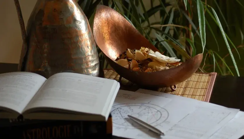 Na stole leży otwarta księga "Astrologie", obok wykresy, długopis i miedziana misa z suszonymi kwiatami. Umiarkowanie tarot uczucia.