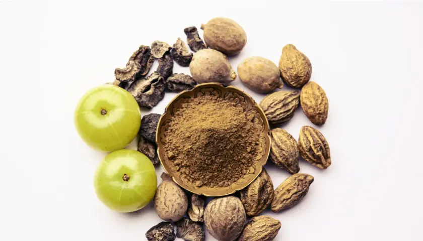 Triphala Pulver in einer Schale, umgeben von Amla-Früchten und getrockneten Haritaki- und Bibhitaki-Samen. Die Triphala Wirkung wird durch diese Zutaten erzielt.